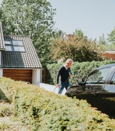 Een man staat op de oprit van zijn huis naast een elektrische auto, en houdt de oplaadstekker ervan in zijn handen.