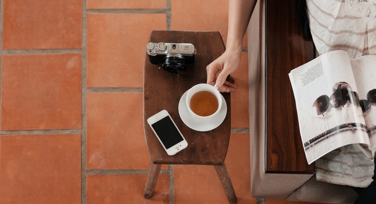 Vrouw zit aan een tafel een tijdschrift te lezen. Ze pakt haar kop koffie van de tafel waar ook een smartphone en een fototoestel liggen. Met de BuitenDeDeurverzekering® van Interpolis kunt u uw spullen buitenshuis in Nederland verzekeren.