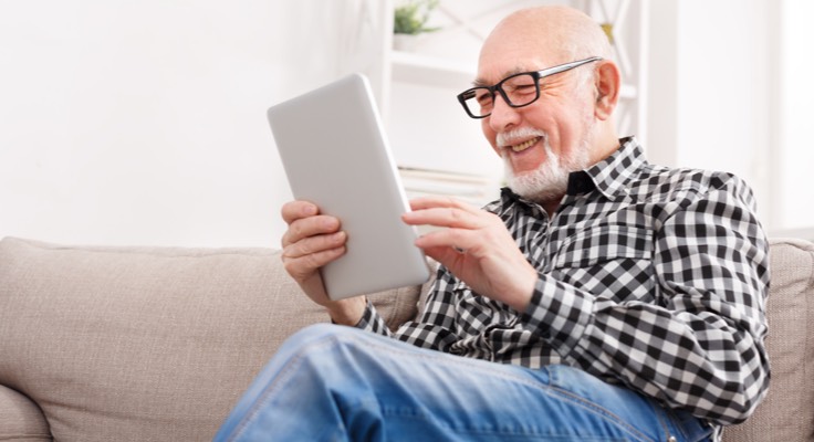 Man zit lachend op de bank met een tablet in zijn hand. Toekomstbestendig wonen, nu en later.