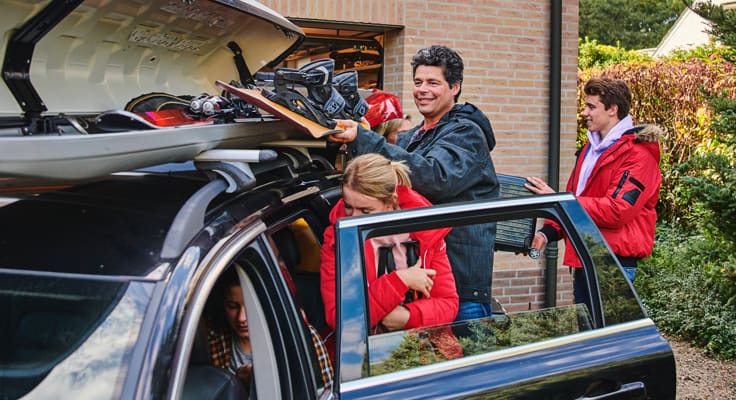 Familie maakt zich klaar om op wintersport te gaan. De dak koffer staat open en de bagage wordt ingeruimd.