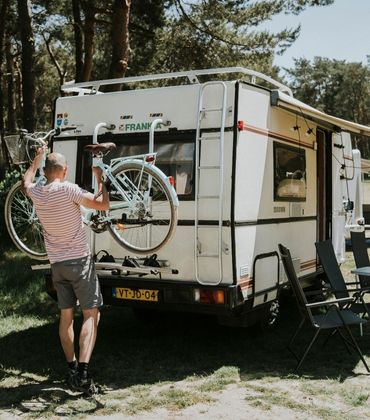 Man staat met fiets in zijn handen achter bij de fietsendrager van de camper en vrouw staat bij luifel met lampjessnoer