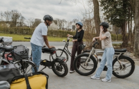 3 jongeren met fietshelm op staan naast hun fietsen en brommer en zijn in gesprek met elkaar.