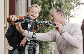 Op de voorgrond fiets een kind in de basisschoolleeftijd geconcentreerd weg, in de richting van de camera. Iets verder staat zijn vader in de deuropening en zwaait het kind uit.