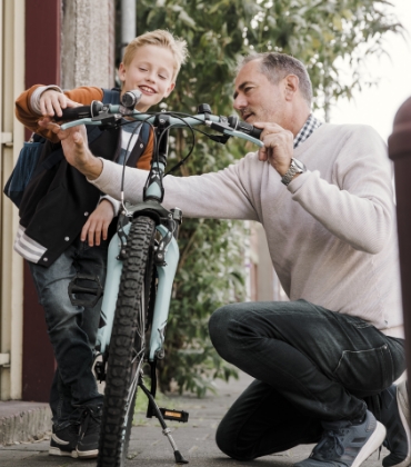 Op de voorgrond fiets een kind in de basisschoolleeftijd geconcentreerd weg, in de richting van de camera. Iets verder staat zijn vader in de deuropening en zwaait het kind uit.