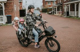 Een jonge vrouw zit op de fiets, met 2 kleine kinderen achterop. Ze hebben aandacht voor hun veiligheid, daarom dragen ze een fietshelm.