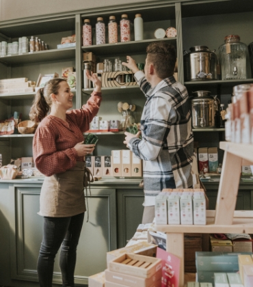 Een verkoopmedewerker en een klant in een sfeervolle winkel reiken beiden met hun hand naar een plank met producten.