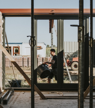 Een man in werkkleding loopt met een speciekuip over een bouwplaats. De foto is gemaakt van binnenuit een pand in aanbouw.
