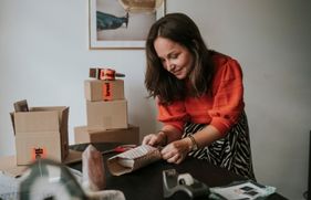 Een vrouw met een rood-oranje trui maakt op een tafel een pakketje verzendklaar. Er staat een aantal verzenddozen op en naast de tafel.