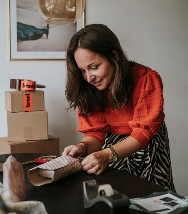 Een vrouw met een rood-oranje trui maakt op een tafel een pakketje verzendklaar. Er staat een aantal verzenddozen op en naast de tafel.