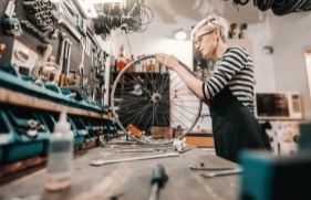 Een vrouw staat in de werkplaats van een fietsenmaker, en inspecteert een fietswiel.