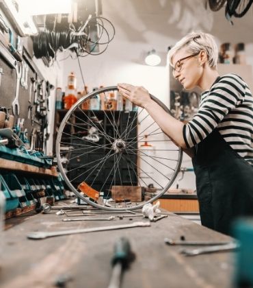 Een vrouw staat in de werkplaats van een fietsenmaker, en inspecteert een fietswiel.