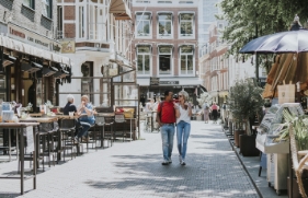 Een man loopt vrolijk met zijn arm om de schouder van een vrouw over straat, langs terrasjes. De zon schijnt. 