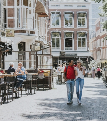 Een man loopt vrolijk met zijn arm om de schouder van een vrouw over straat, langs terrasjes. De zon schijnt. 