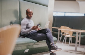 Een man zit op een groene bank, met zijn laptop op schoot en zijn smartphone in de hand. Hij kijkt op het scherm van zijn telefoon.