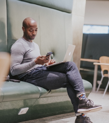 Een man zit op een groene bank, met zijn laptop op schoot en zijn smartphone in de hand. Hij kijkt op het scherm van zijn telefoon.