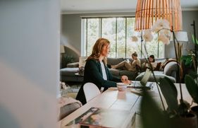 Vrouw werkt aan eetkamertafel en dochters zitten op de bank