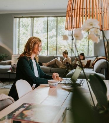 Vrouw werkt aan eetkamertafel en dochters zitten op de bank