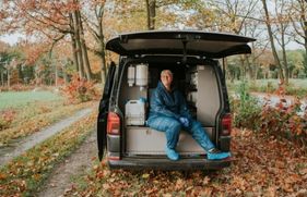 Dierenarts Janny Hermans zit ontspannen op de achterdorpel van een bedrijfswagen, die in een bosrijke omgeving geparkeerd staat.