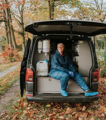 Dierenarts Janny Hermans zit ontspannen op de achterdorpel van een bedrijfswagen, die in een bosrijke omgeving geparkeerd staat.