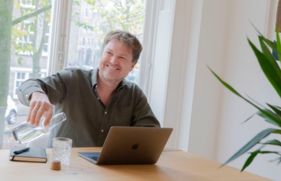 Guido Heezen zit achter zijn laptop en schenkt water in een drinkglas vanuit een flesje.
