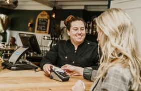 Een klant van een horeca-gelegenheid rekent met haar pinpas af bij een vrolijk lachende medewerker.