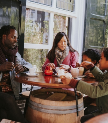Een man, vrouw en 2 kinderen zitten met shawls en mutsen buiten aan een ronde tafel met koffie en chocomel.