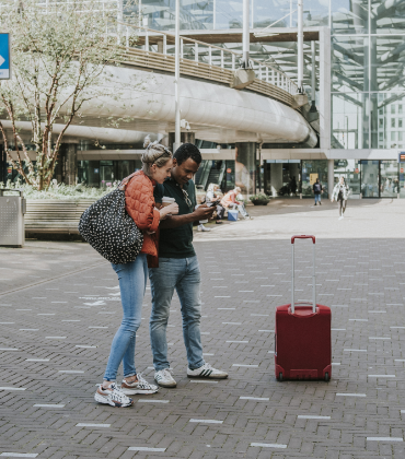 Man en vrouw staan met koffie in de hand en bagage op een stationsplein en kijken op de telefoon die de man vasthoudt