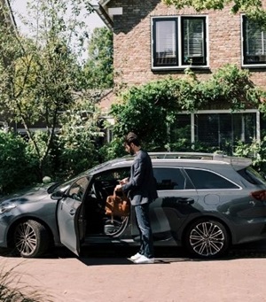 Een man staat naast zijn auto met geopend portier. Hij heeft zijn tas en telefoon in de hand, en is klaar om in te stappen.