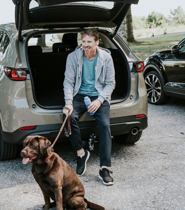 Man zit achter op de rand van de auto met de achterklep open en heeft een hond vast