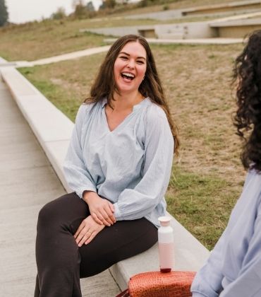 Een vrouw zit op de betonnen rand van een perk en lacht uitbundig naar een andere vrouw, met wie ze in gesprek is.