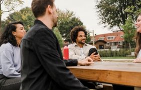 4 jonge mensen zitten aan tafel in een park en kletsen en lachen