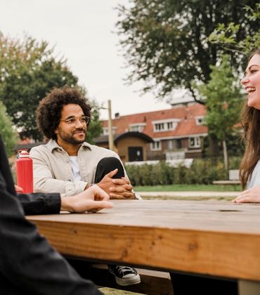 4 jonge mensen zitten aan tafel in een park en kletsen en lachen