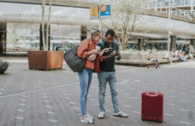 Een stel staat op een stationsplein aan het begin van een reis. Ze kijken samen naar het scherm van een mobiele telefoon ter voorbereiding op hun reis.