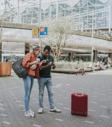 Een stel staat op een stationsplein aan het begin van een reis. Ze kijken samen naar het scherm van een mobiele telefoon ter voorbereiding op hun reis.