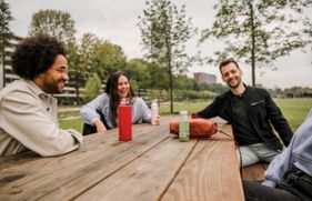 Een groepje jonge mensen zit bij elkaar aan een houten picknicktafel in een stadspark.