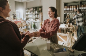 Een winkelier geeft een ingepakt doosje aan een klant, terwijl ze allebei vrolijk kijken.