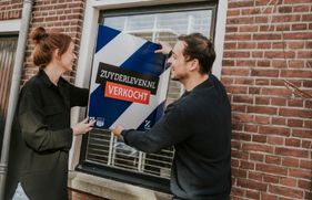 Man en vrouw lachen en houden een verkocht bord vast bij het raam van een woning