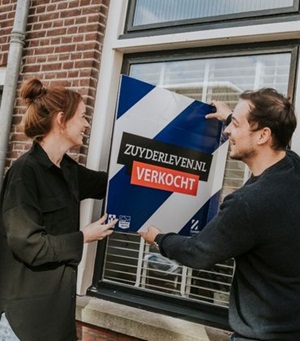 Man en vrouw lachen en houden een verkocht bord vast bij het raam van een woning