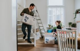 Vrouw zit op de vloer naast een trap. Man loopt met een verhuisdoos in zijn handen.