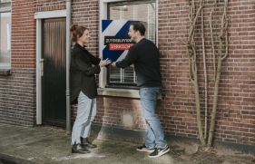 Een man en vrouw staan bij het raam van een woning en houden het verkocht bord vast