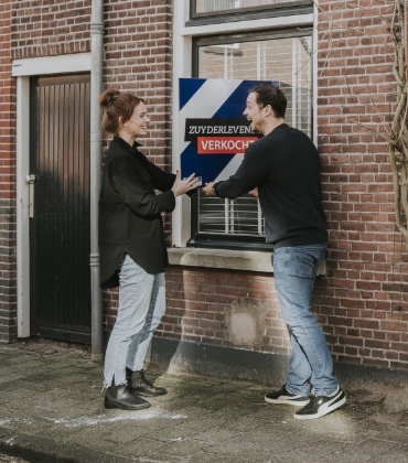 Een man en vrouw staan bij het raam van een woning en houden het verkocht bord vast