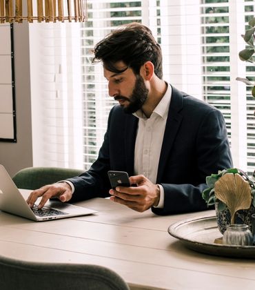 Man zit aan tafel achter zijn laptop en heeft een telefoon in zijn hand