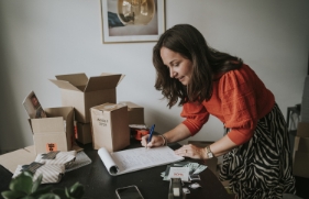 Een glimlachende vrouw met een rood-oranje trui buigt zich in een thuiswerkomgeving over haar werkplek. Ze schrijft iets op een papieren schrijfblok. Naast haar op het bureau staan enkele kartonnen dozen.