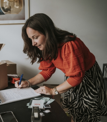 Een glimlachende vrouw met een rood-oranje trui buigt zich in een thuiswerkomgeving over haar werkplek. Ze schrijft iets op een papieren schrijfblok. Naast haar op het bureau staan enkele kartonnen dozen.