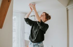 Een vrouw controleert of bevestigt een rookmelder aan het plafond. Het is belangrijk om regelmatig je rookmelders te testen, schoon te maken en te onderhouden om zeker te weten dat deze je op tijd waarschuwen in geval van rook of brand