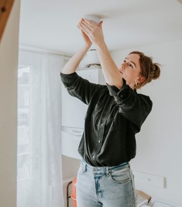 Een vrouw controleert of bevestigt een rookmelder aan het plafond. Het is belangrijk om regelmatig je rookmelders te testen, schoon te maken en te onderhouden om zeker te weten dat deze je op tijd waarschuwen in geval van rook of brand