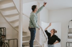 Man staat op trap en houdt een rookmelder tegen het plafond en vrouw kijkt naar de rookmelder