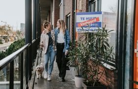 2 vrouwen lopen met een aangelijnde hond over de galerij van een appartementencomplex. Ze kijken elkaar aan en hebben een lach op hun gezicht. Op het appartement waar ze langslopen, hangt een bord met 'Verhuurd' aan het raam.