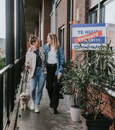 2 vrouwen lopen met een aangelijnde hond over de galerij van een appartementencomplex. Ze kijken elkaar aan en hebben een lach op hun gezicht. Op het appartement waar ze langslopen, hangt een bord met 'Verhuurd' aan het raam.