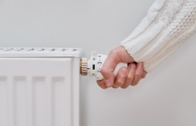 Iemand pakt met 1 hand de thermostaatknop van de radiator vast, om de verwarming hoger of lager te kunnen zetten.